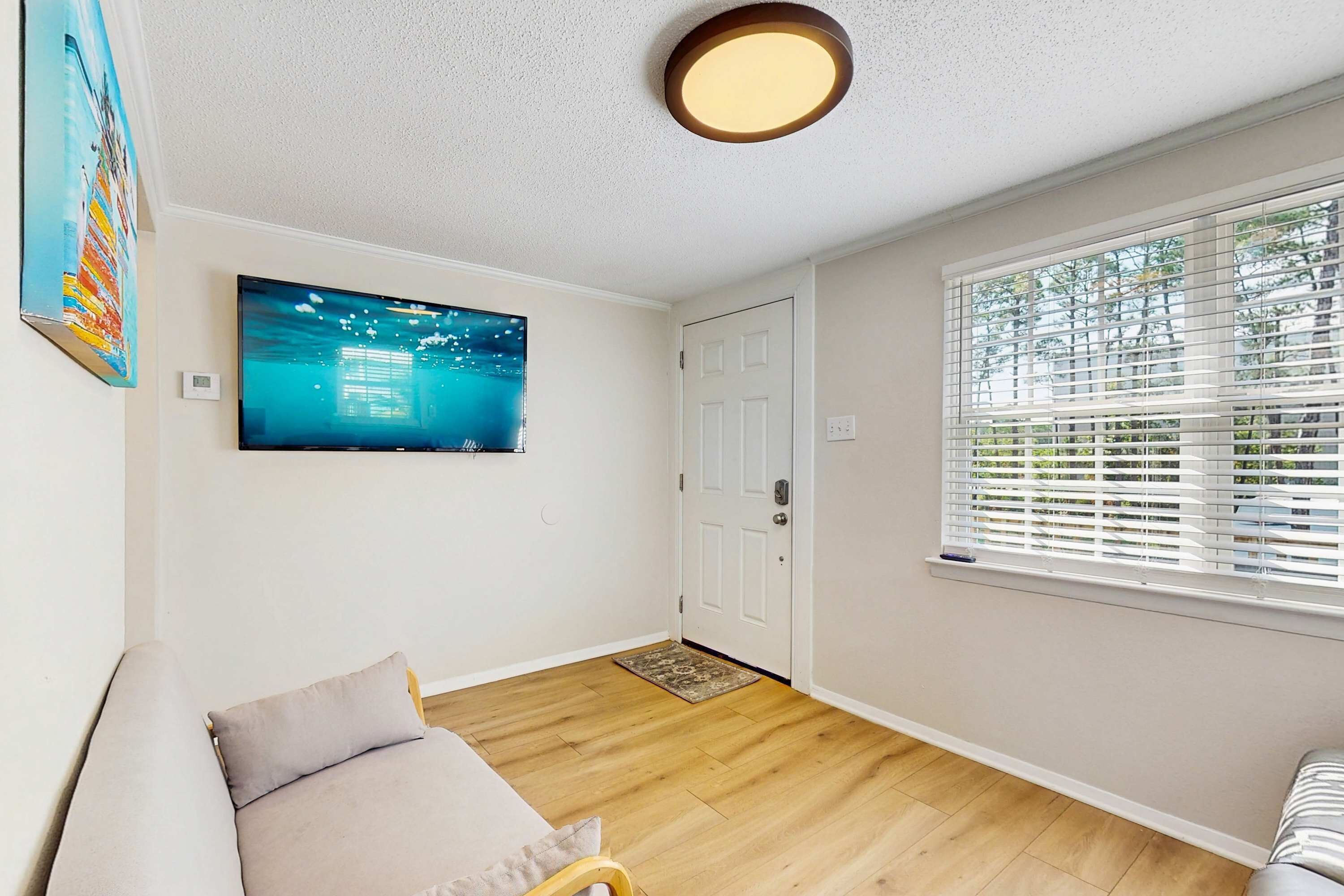 A room with a flat-screen TV mounted on the wall, a door, a window with blinds, a wooden floor, and a sofa.