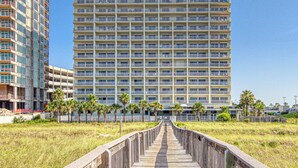 A multi-story hotel with a central atrium, a boardwalk leading to the entrance, and palm trees lining the walkway. - Updated Gulf front condo w/ shared pool, hot tub, and sauna, & beach access (Orange Beach)