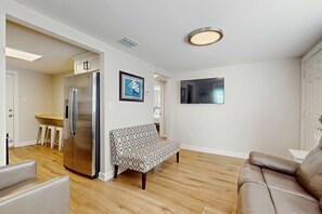 A modern living room with a sofa, a brown leather couch, a patterned sofa, a kitchen area with a stainless steel refrigerator, wooden flooring, a flat-screen TV mounted on the wall, and a ceiling light fixture. - Two charming homes with sparkling water views and close to the beach (Gulf Shores)