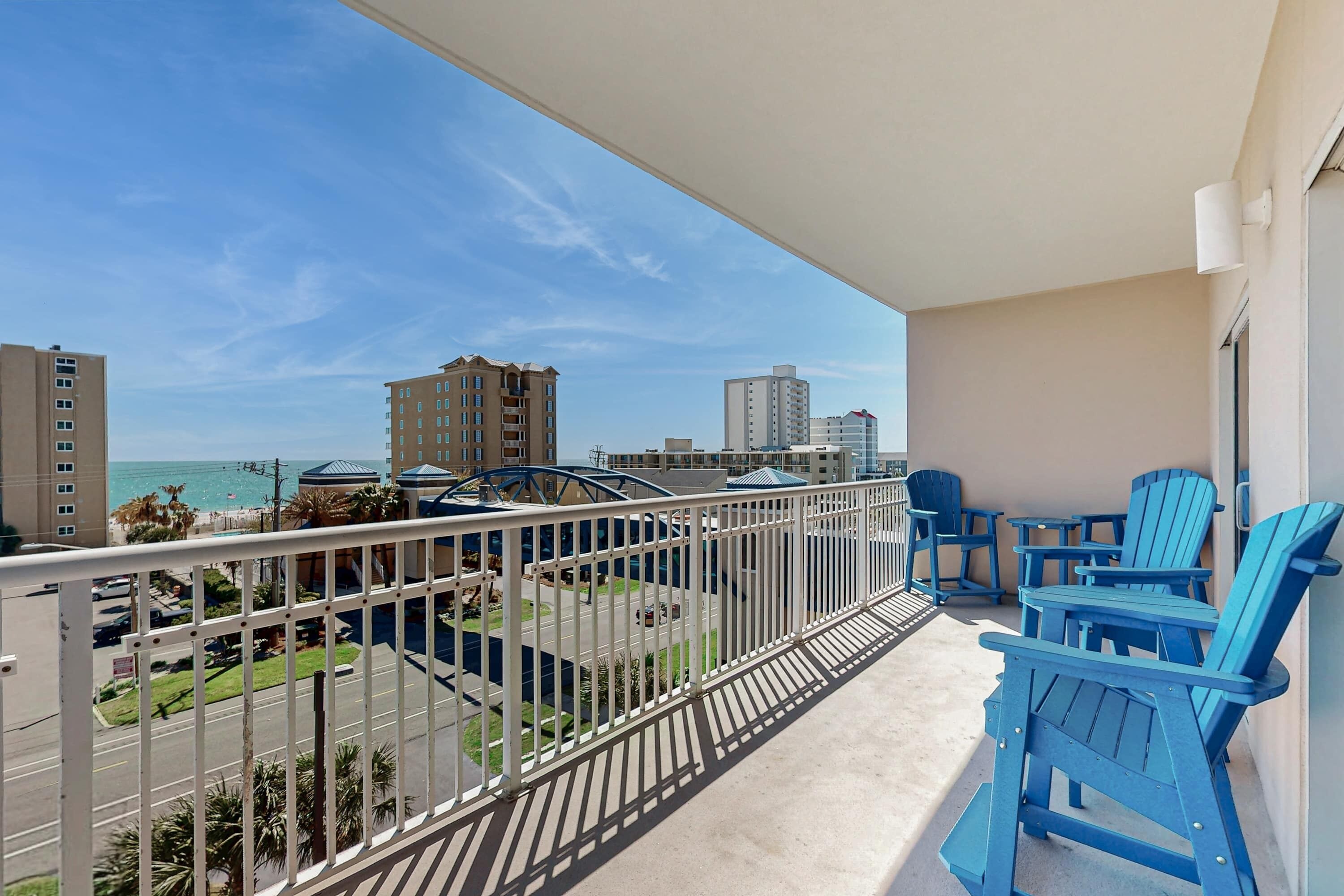 A balcony with a view of the beach and cityscape, featuring blue Adirondack chairs and a railing.