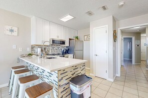 Eine moderne Küche mit weißen Schränken, einer zentralen Kochinsel mit Hockern, einer Mikrowelle, einem Kühlschrank und einer gefliesten Spritzschutzwand.