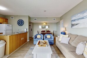 A living room with a kitchen, a clock, a dining area, and a couch. - Bright coastal condo with Gulf views, pools, hot tub & private balcony (Gulf Shores)
