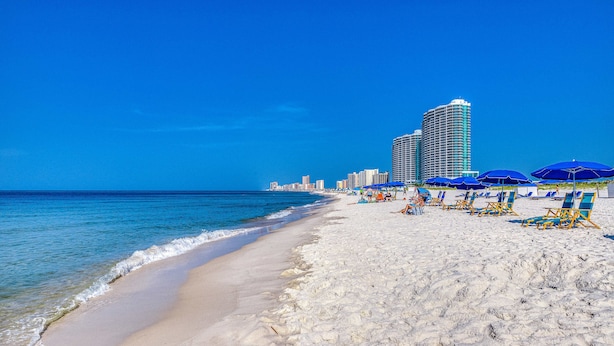 Una spiaggia con sabbia bianca, un cielo azzurro intenso e un alto edificio di un albergo sullo sfondo.
