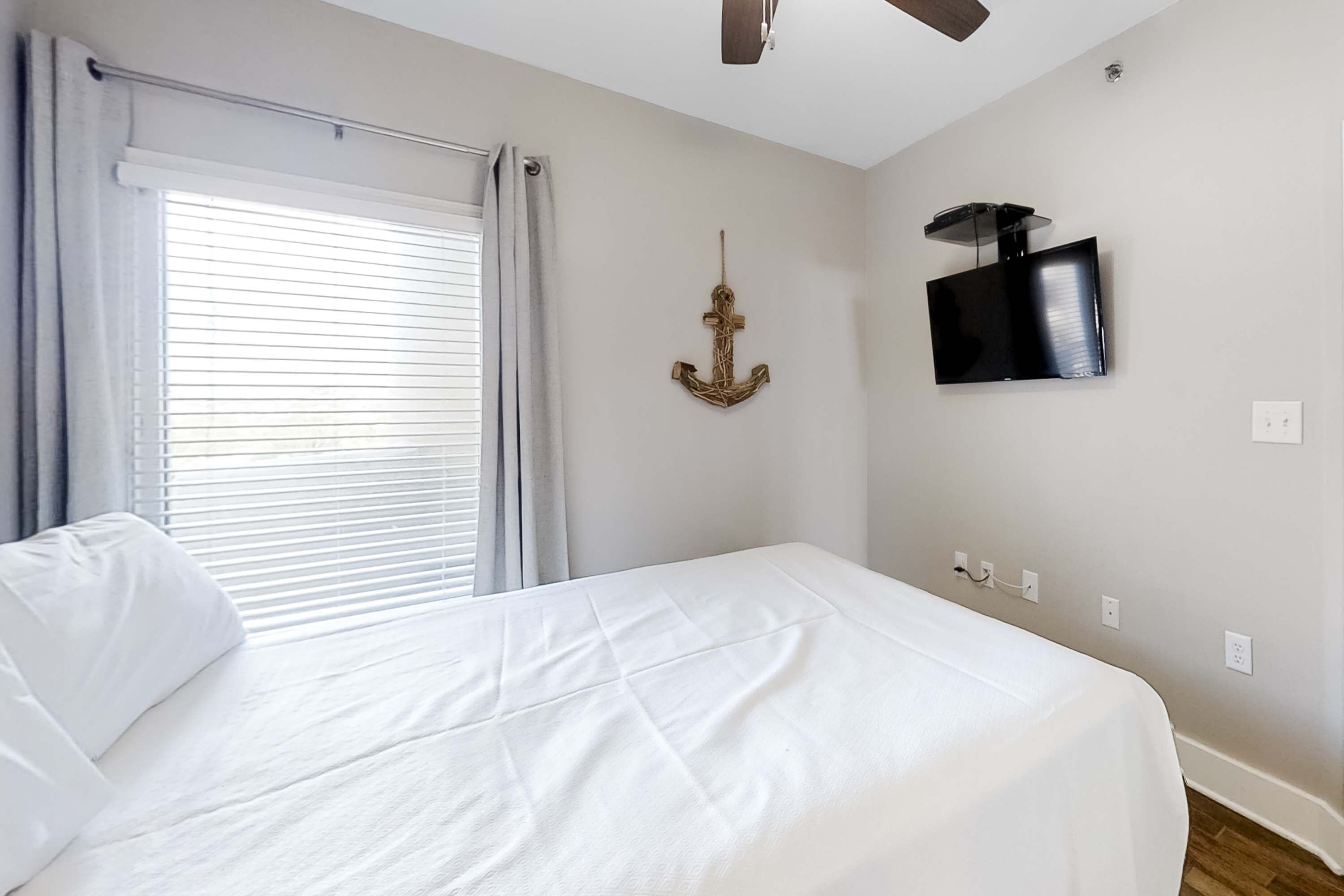 A bedroom with a bed, a window with curtains, a wall-mounted TV, and a ceiling fan.