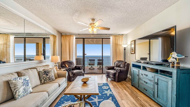 Een ruime woonkamer met een bank, fauteuils, een salontafel en een televisie. Er is een balkon met uitzicht op de oceaan.