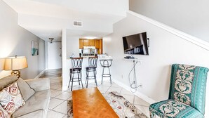A living room with a sofa, a wooden coffee table, a patterned rug, a TV mounted on the wall, and a kitchen area in the background. - Bright resort condo with pool access, cozy patio, plus a washer/dryer (Orange Beach)