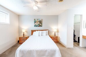 A bedroom with a bed, two bedside tables with lamps, a ceiling fan, a painting on the wall, and a doorway leading to another room.
