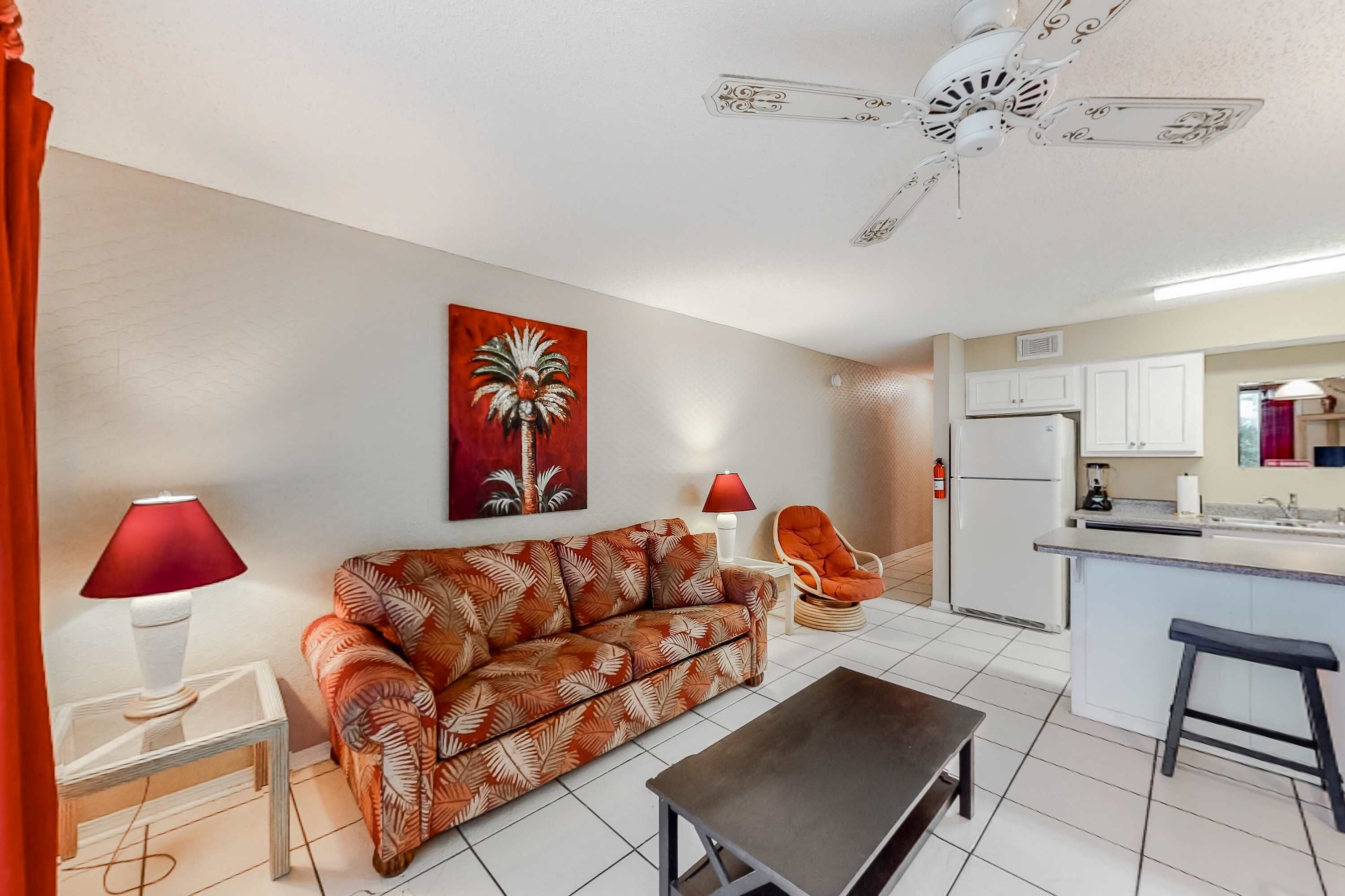 A living room with a sofa, a coffee table, a red lamp, a chair, and a kitchen area with cabinets and a refrigerator.