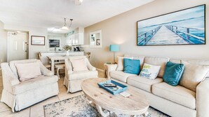 A living room with a sofa, chairs, a coffee table, a painting of a pier, and a ceiling fan. - Beautiful home in beachfront luxury complex with pool, sauna & WiFi (Orange Beach)