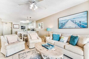 A living room with a sofa, chairs, a coffee table, a painting of a pier, and a ceiling fan. - Beautiful home in beachfront luxury complex with pool, sauna & WiFi (Orange Beach)