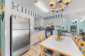 A modern kitchen with a stainless steel refrigerator, white cabinets, and a dining area with a white table and wooden chairs. - Splendid beachfront condo with pool, tennis court, sauna, & WiFi (Orange Beach)