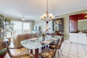 A dining area with a glass table, wicker chairs, a chandelier, a potted plant, and a view of the outdoors. - 9th-floor, Gulf-front condo with balcony, pools, hot tubs, gym & beach access (Orange Beach)