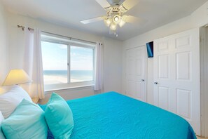 Ein Schlafzimmer mit Bett, Deckenventilator, Fenster mit Vorhängen und Blick auf den Strand.