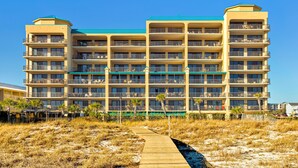 Un hôtel à plusieurs étages doté de balcons, offrant une vue sur la plage et un chemin de sable menant à l’entrée.