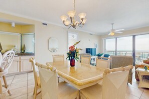 A dining area with a large table, chairs, a chandelier, and a view of the ocean through large windows. - 5th-floor beachfront condo with balcony, wet bar, pool, hot tub, sauna & gym (Orange Beach)