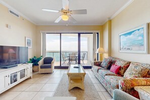 A living room with a television, sofa, coffee table, and a balcony with a view. - 5th-floor beachfront condo with balcony, wet bar, pool, hot tub, sauna & gym (Orange Beach)