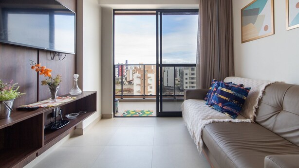 A modern living room with a sofa, a flat-screen TV, a balcony with a city view, and a small table with decorative items. - Park Del Prado #603 - Intermares by Carpediem (Cabedelo)