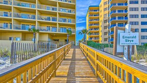 Eine Holzpromenade führt zu einem Hotel mit mehreren Balkonen und einem Schild mit der Aufschrift 'Emerald Skye 26034 Perdido Beach Blvd.'