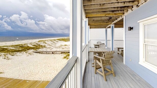 Una terraza de madera con vista a la playa y al océano, una mesa con sillas y un área cubierta con barandilla.