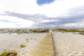 Ein hölzerner Steg führt durch eine sandige Küstenlandschaft mit Dünen im Hintergrund.