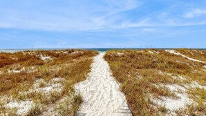 Ein sandiger Pfad führt zu einem Strand, der von Dünen gesäumt ist.