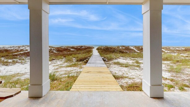 Ein Holzsteg führt zu einem Strand mit weißen Säulen und blauem Himmel.