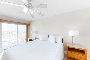 A hotel room with a large bed, a ceiling fan, a nightstand with a lamp, and a view of the beach. - Spacious beachfront wedding-friendly home with multiple decks & shared pool (Orange Beach)
