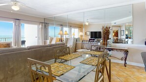 Una sala de estar con una mesa de comedor de vidrio, asientos acolchados, un ventilador de techo y vista al océano.