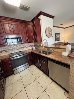 A kitchen with dark wooden cabinets, stainless steel appliances, a granite countertop, and a tiled floor. - Ground-Floor Condo near the Beach w/Private Patio, Shared Pool, and Central A/C (Orange Beach)