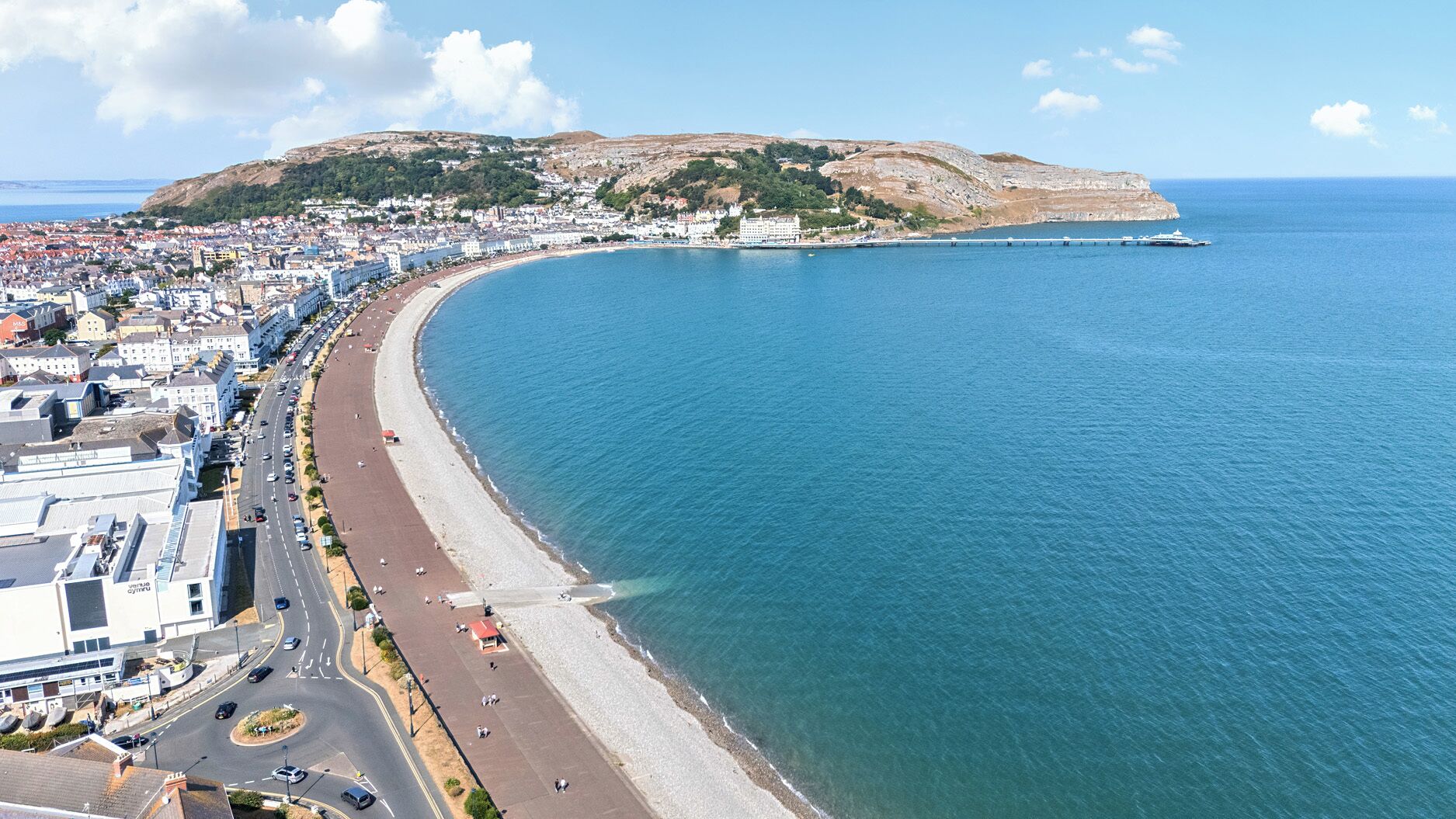 Paisagem costeira com uma praia curva, uma colina imponente e uma ponte ligando a terra ao mar.
