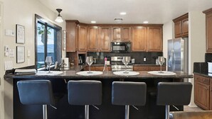 Una cocina moderna con armarios de madera, una isla central con taburetes de bar, electrodomésticos de acero inoxidable y vistas a través del ventanal.