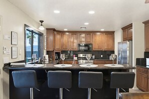 A modern kitchen with wooden cabinets, a central island with bar stools, stainless steel appliances, and a view through the window. - Havasu Pool Home | Spa, Games & Boat Parking (Lake Havasu City)
