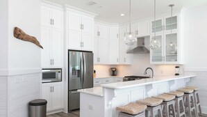 A modern kitchen with white cabinetry, stainless steel appliances, and a central island with bar stools. - Luxury Gulf-front home w private splash pool & direct beach access (Orange Beach)