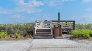 Een houten looppad dat leidt naar een houten gebouw met schoorsteen, omgeven door hoog gras en een heldere lucht.