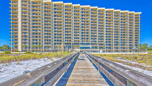 A large hotel with multiple floors, a wooden boardwalk leading to the entrance, and a clear blue sky. - Waterfront condo w/ balcony, multiple pools, lighted tennis courts, hot tub (Orange Beach)
