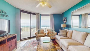A living room with a TV, sofa, chairs, a coffee table, and a ceiling fan. - Waterfront condo w/ balcony, multiple pools, lighted tennis courts, hot tub (Orange Beach)