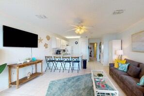 A living room with a sofa, a coffee table, a TV, and a dining area with chairs and a table. - Beautiful oceanfront condo with pool, hot tub, beach access, balcony, & WiFi (Gulf Shores)