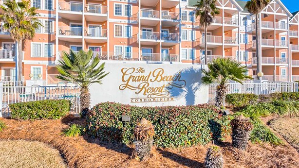 Een meerlaags gebouw met balkons, een bord waarop 'The Grand Beach Resort Condominium' staat, en aangelegde gebieden met palmbomen en struiken.