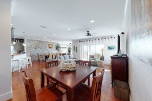 A spacious living area with a dining table, chairs, a kitchen in the background, and a large window. - Delightful escape with direct beach access, WiFi, furnished deck, & full kitchen (Gulf Shores)