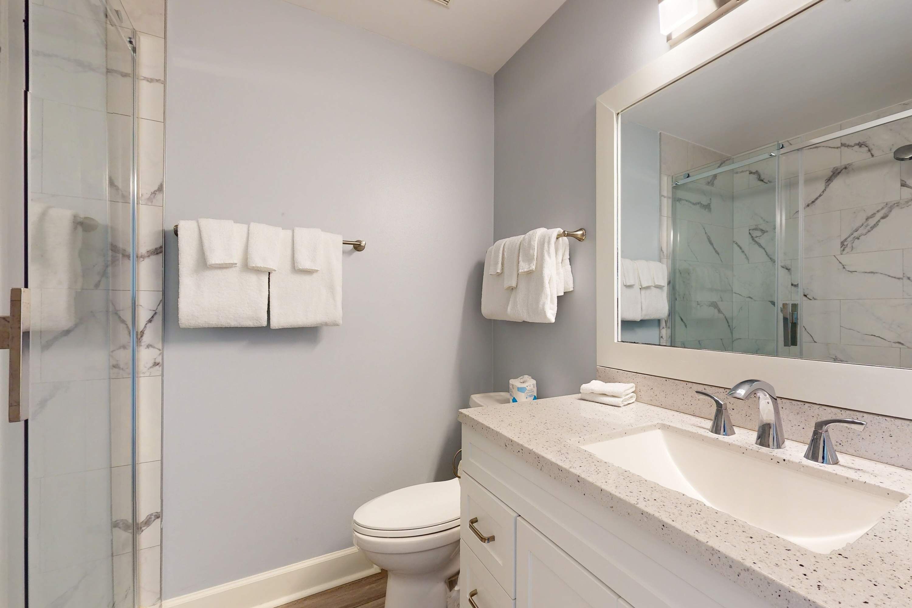 A bathroom with a glass shower enclosure, a white toilet, a sink with a large mirror, and towels on the wall.