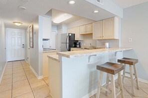A kitchen with a central island, bar stools, a refrigerator, and a coffee maker. - Boardwalk 283 gulf-view condo with WiFi, central ac, balcony, & outdoor pool (Gulf Shores)