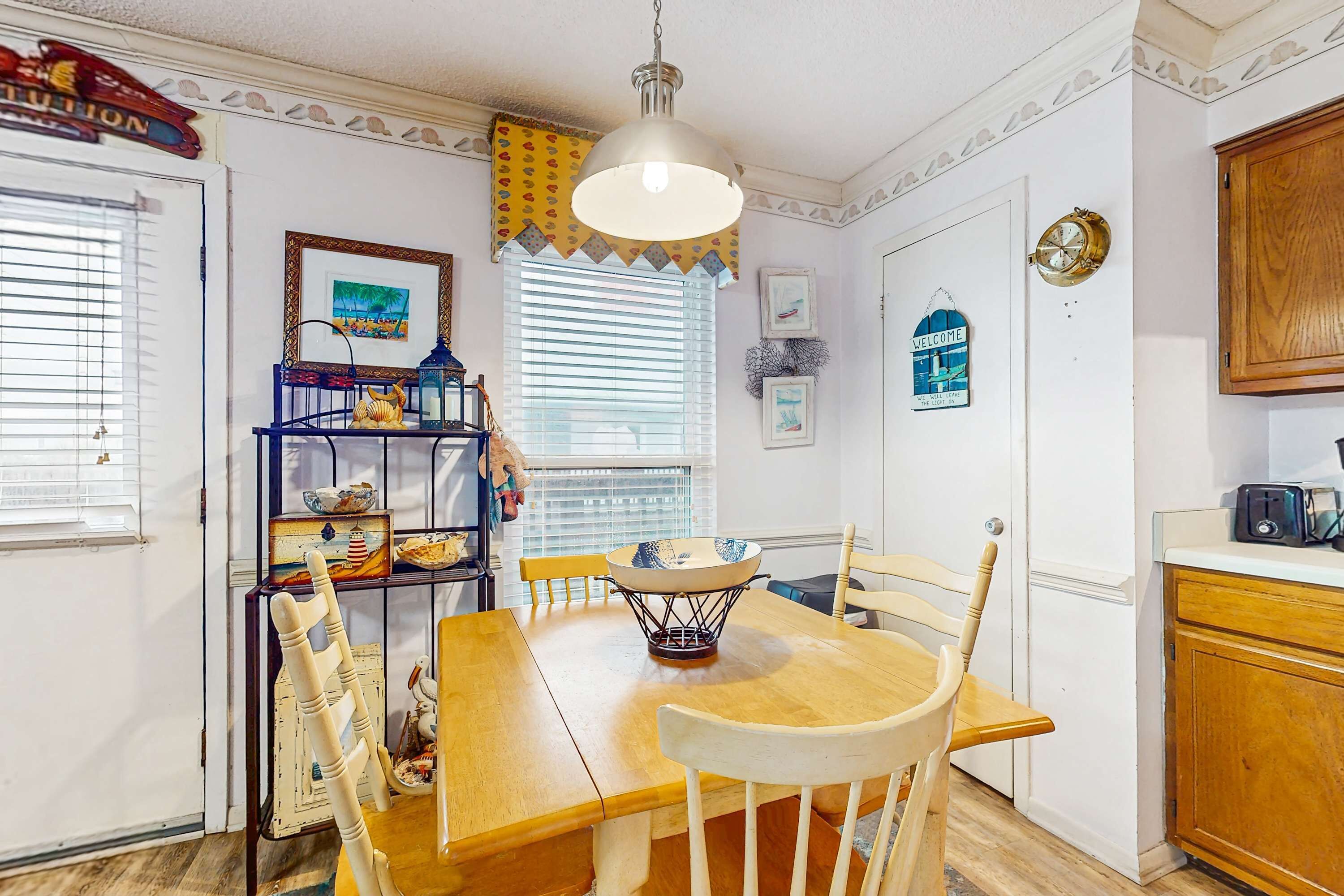 A dining area with a wooden table, chairs, a hanging light fixture, and a window with blinds.