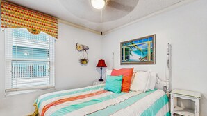 Un dormitorio con cama, ventana con persianas, lámpara de pared, cuadro enmarcado y una pequeña mesa.