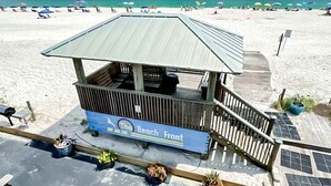 Un quiosco frente a la playa llamado 'The Beach Front', con techo de metal, terraza de madera y plantas en macetas.