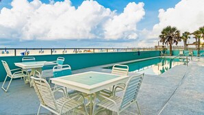 Uma área junto à piscina com mesas e cadeiras, uma piscina e vista para a praia.