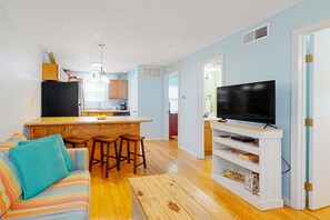 A living room with a sofa, a coffee table, a TV on a stand, a kitchen area with a fridge, and a dining table with chairs. - Gorgeous Condo in Beachfront Location w/Shared Pool, Central AC, Free WiFi (Gulf Shores)