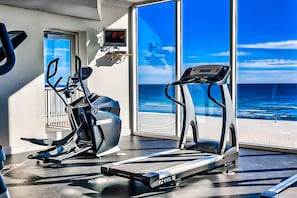 A fitness area with treadmills and an elliptical machine, large windows with a view of the ocean, and a television mounted on the wall. - Gulf-front penthouse with pool, sauna, washer,dryer, & central AC (Gulf Shores)