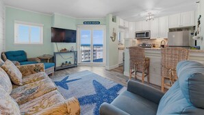 Ein Wohnzimmer mit einer Couch, einem Couchtisch und einem Teppich mit Sternenmuster. Es gibt eine Küchenzeile mit weißen Schränken, einer Mikrowelle und einem Kühlschrank. Ein Schild mit der Aufschrift 'BEACH HOUSE' ist sichtbar.