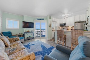 Ett vardagsrum med en soffa, ett soffbord och en matta med stjärnmönster. Det finns ett köksområde med vita skåp, en mikrovågsugn och ett kylskåp. En skylt med texten ”STRANDHUS” syns.