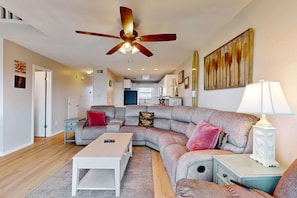 A living room with a sectional sofa, a coffee table, a side table, a ceiling fan, and a kitchen in the background. - Multi-Level, Beachfront Condo w/ Gulf Views, Deck, WiFi, AC & Shared Pool (Gulf Shores)